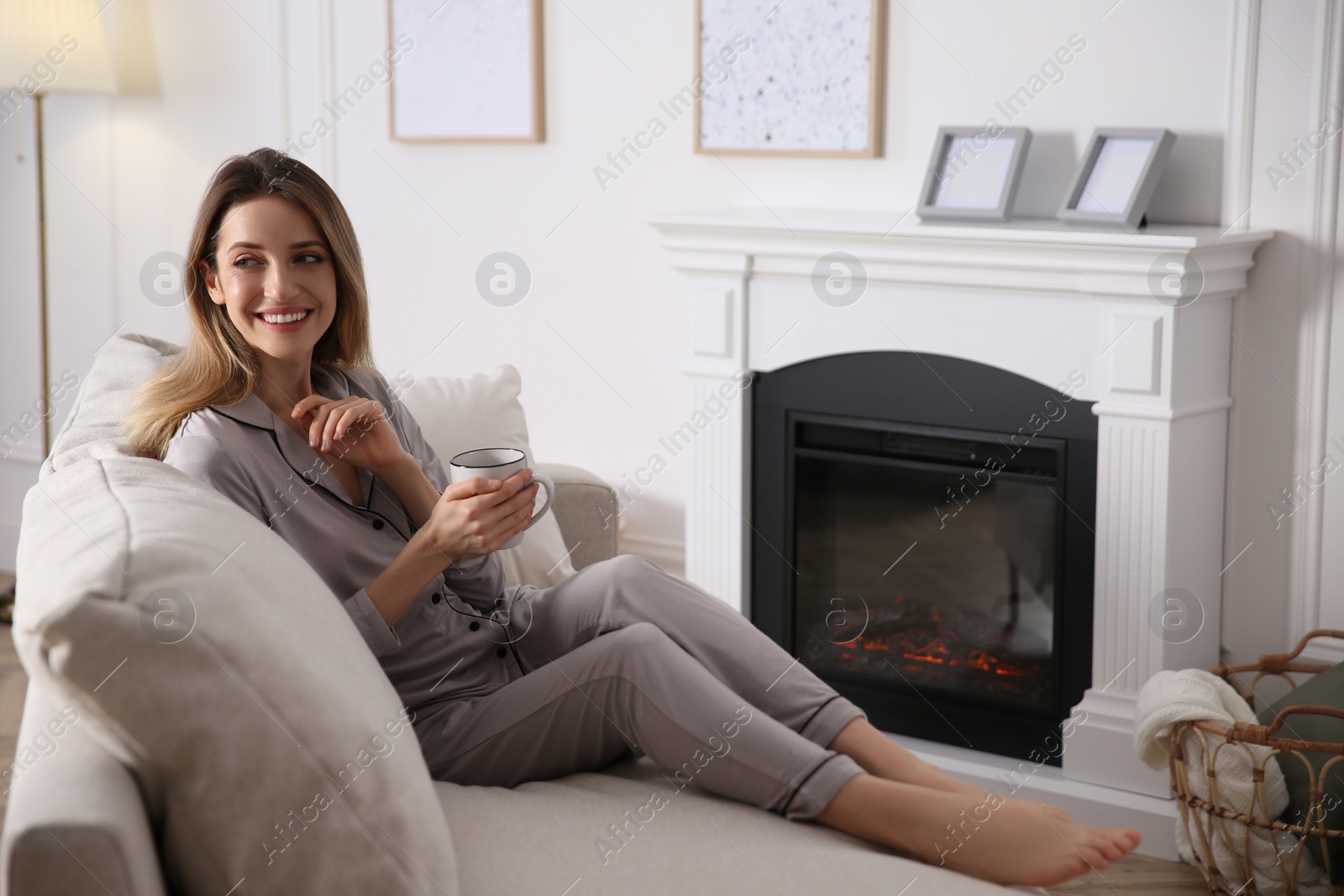 Photo of Young woman with cup of hot drink resting on sofa near fireplace at home