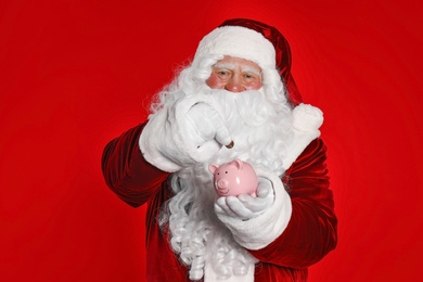Photo of Santa Claus putting coin into piggy bank on red background