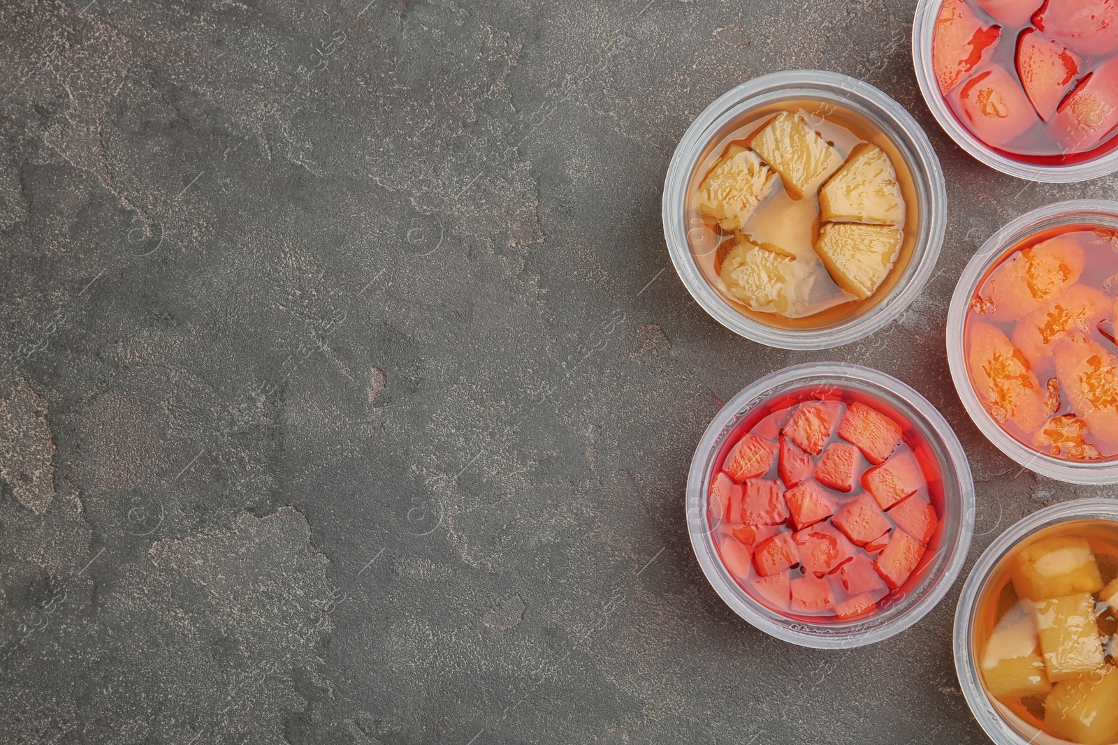 Photo of Plastic containers of tasty jelly desserts with fruit pieces on grey background, flat lay. Space for text