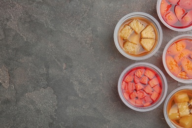 Plastic containers of tasty jelly desserts with fruit pieces on grey background, flat lay. Space for text