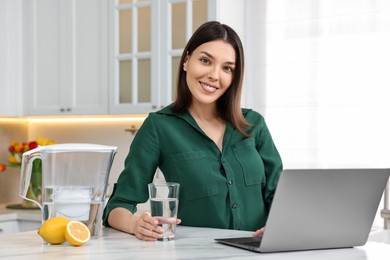 Woman with glass of water and filter jug near laptop in kitchen