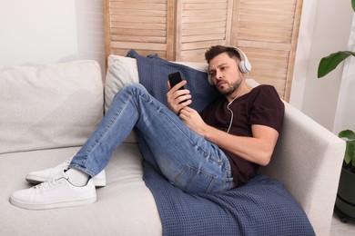 Upset man listening to music through headphones on sofa at home. Loneliness concept