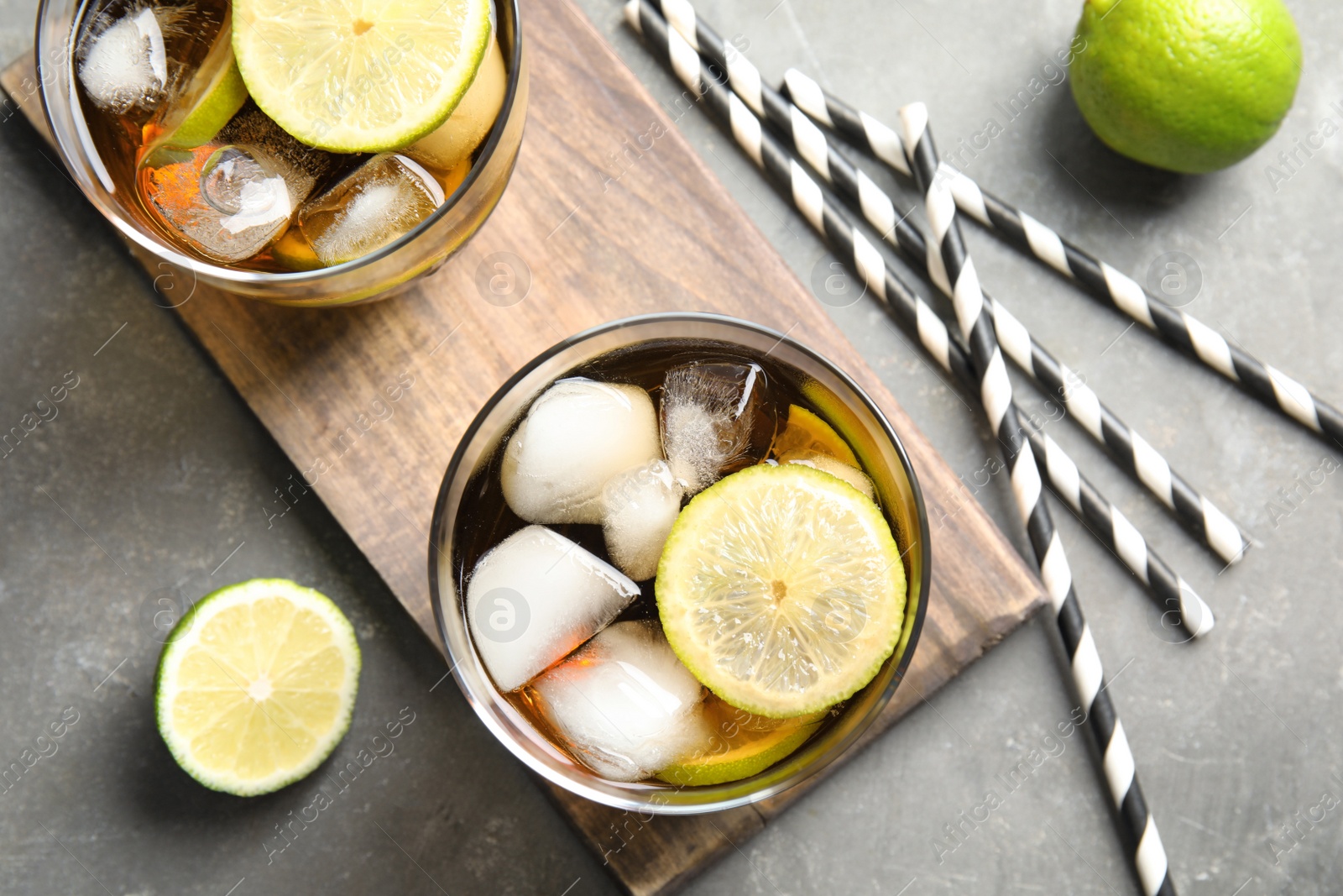 Photo of Glasses of cocktail with cola, ice and cut lime on grey background, flat lay