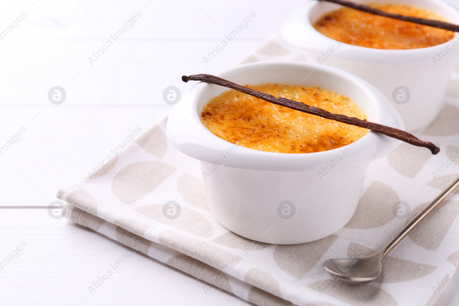 Photo of Delicious creme brulee in bowls, vanilla pods and spoon on white wooden table, closeup