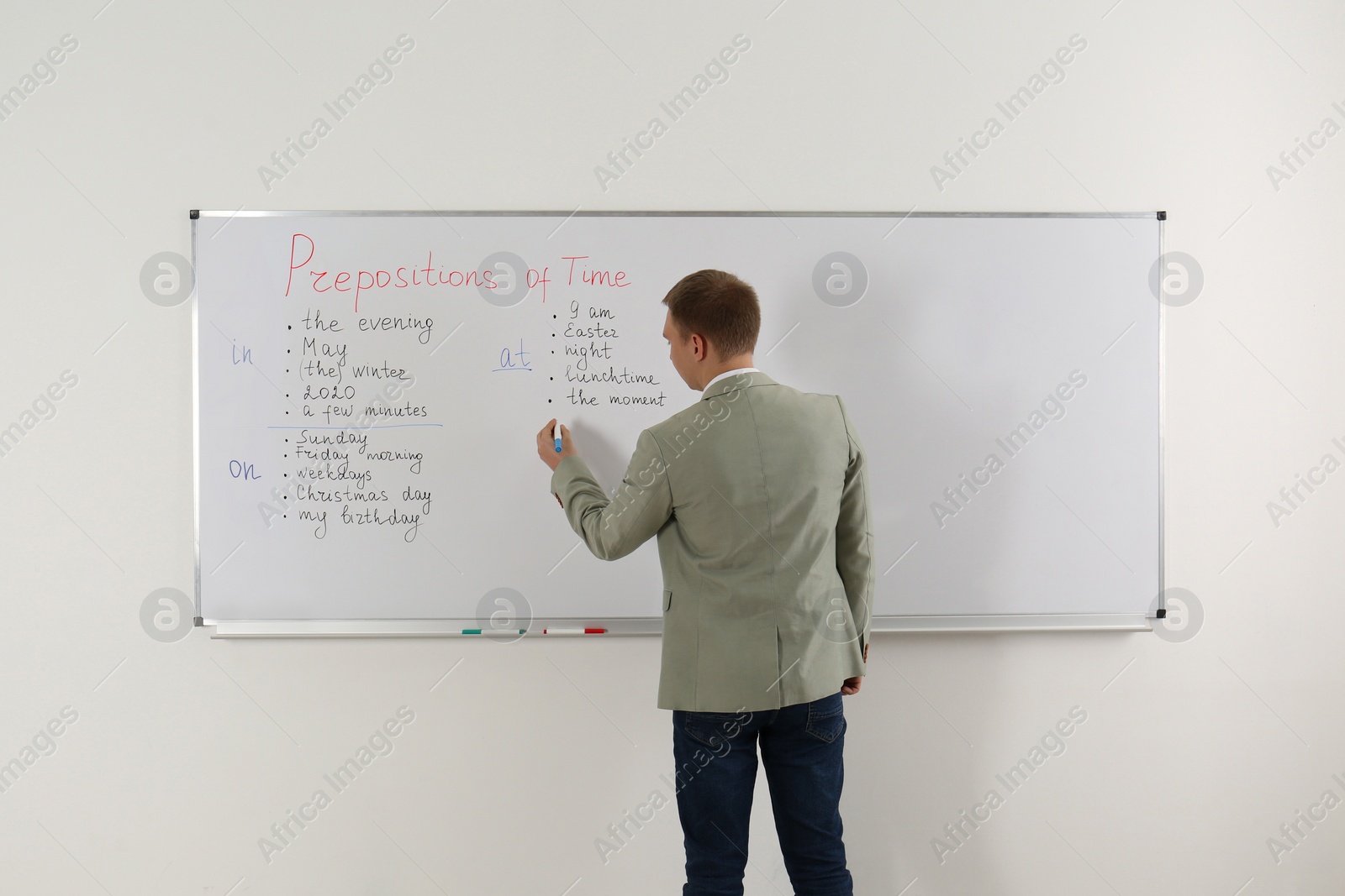 Photo of English teacher giving lesson on prepositions of time near whiteboard in classroom, back view