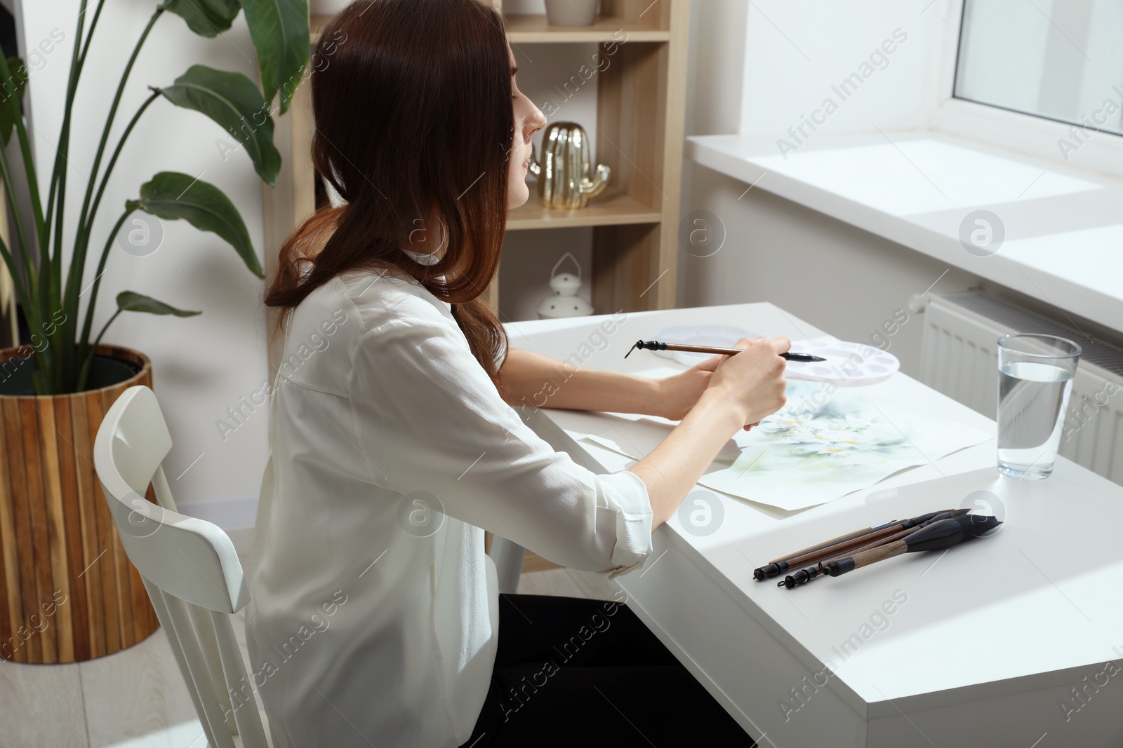 Photo of Woman painting flowers with watercolor at white table in room. Creative artwork