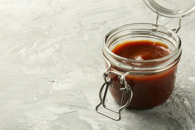 Photo of Tasty barbeque sauce in jar on grey textured table, closeup. Space for text