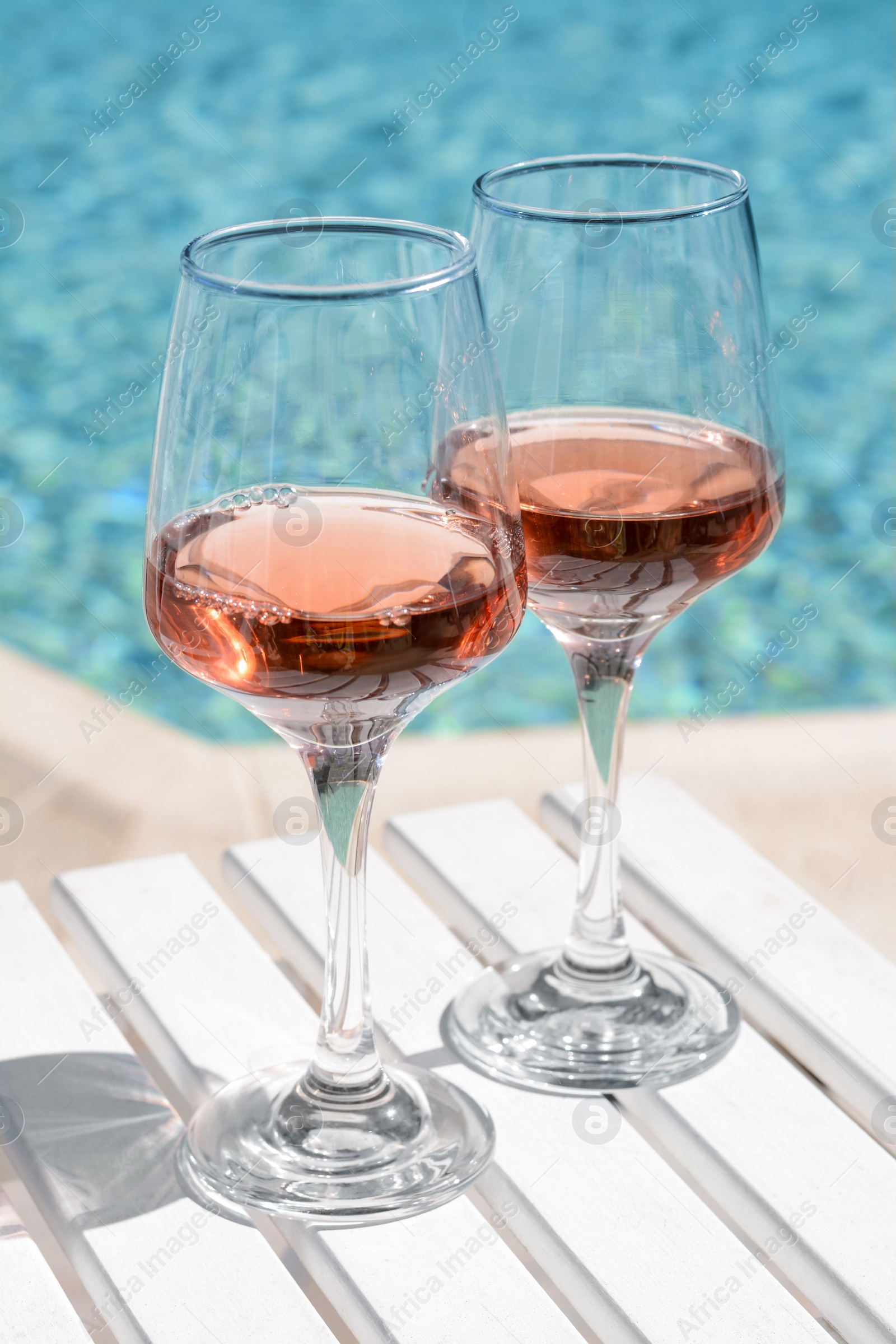 Photo of Glasses of tasty rose wine on white wooden table near swimming pool, closeup