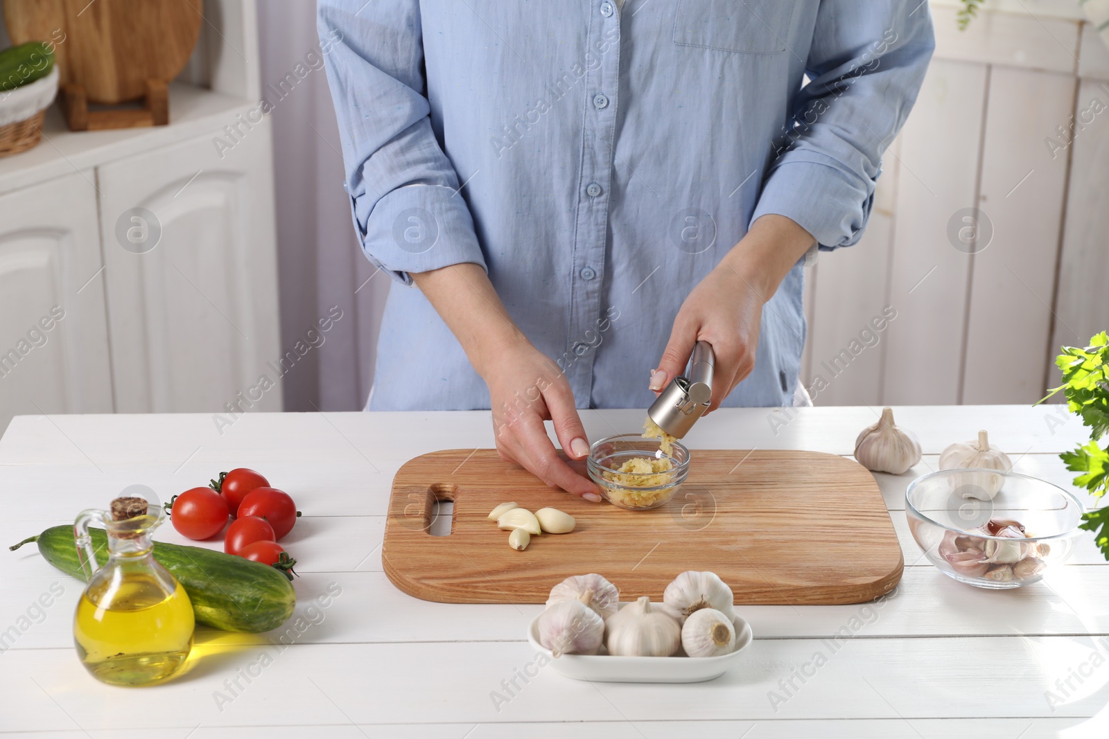 Photo of Woman squeezing garlic with press at white wooden table in kitchen, closeup