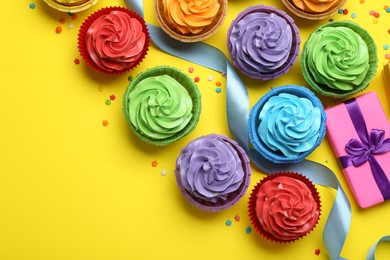 Photo of Delicious birthday cupcakes, gift, ribbon and sprinkles on yellow background, flat lay. Space for text