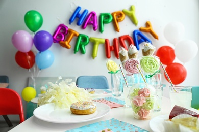 Photo of Table with treats and phrase HAPPY BIRTHDAY made of colorful balloon letters in living room