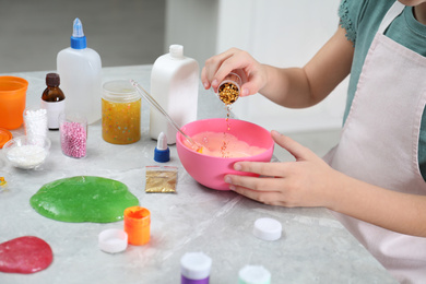 Photo of Little girl adding colored sparkles into homemade slime toy at table, closeup of hands