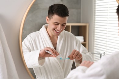 Photo of Happy man squeezing toothpaste from tube onto toothbrush near mirror in bathroom