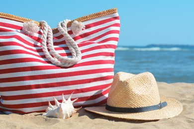 Photo of Stylish striped bag with straw hat and seashell on sandy beach near sea