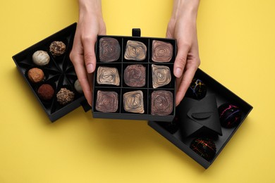 Photo of Woman holding box of delicious chocolate candies on yellow background, top view