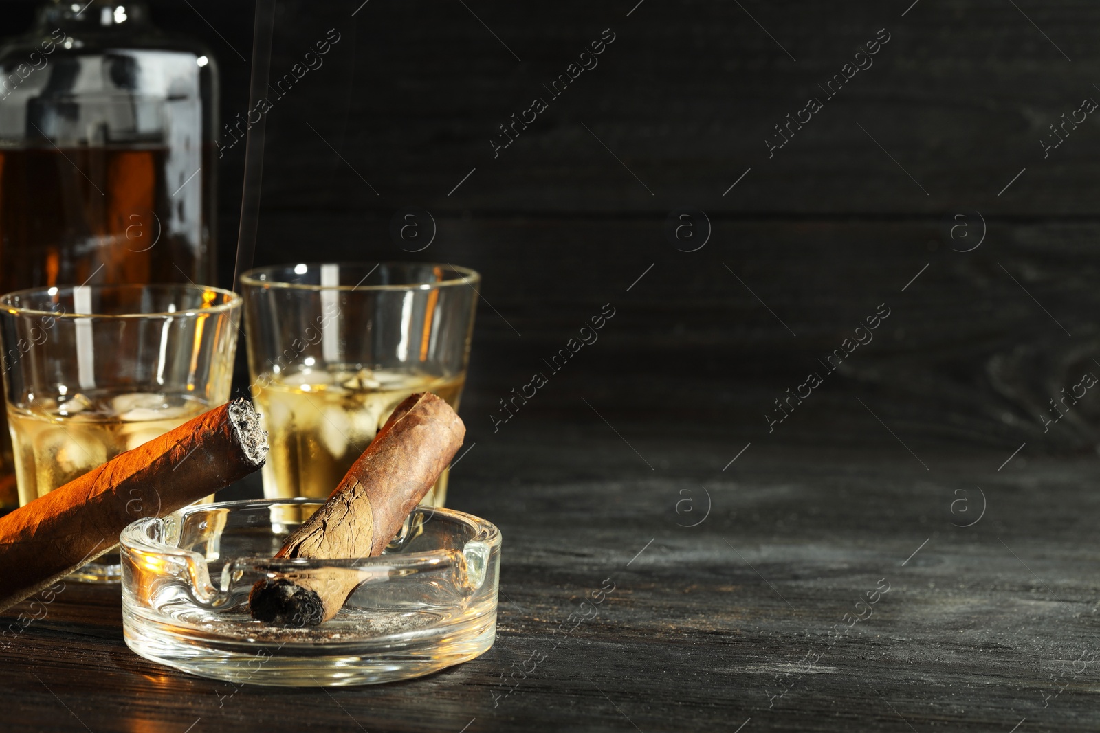 Photo of Cigars, ashtray and whiskey with ice cubes on black wooden table. Space for text