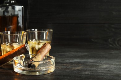 Cigars, ashtray and whiskey with ice cubes on black wooden table. Space for text