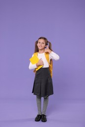 Photo of Smiling schoolgirl with backpack and book showing thumbs up on violet background