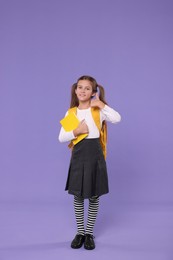 Smiling schoolgirl with backpack and book showing thumbs up on violet background