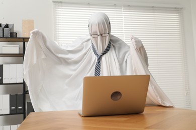 Photo of Overworked ghost. Man covered with white sheet using laptop at wooden table in office