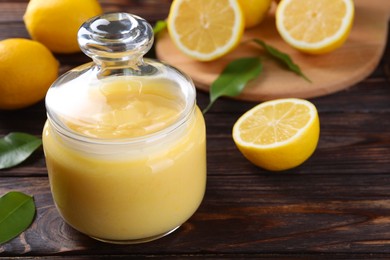 Photo of Delicious lemon curd in glass jar, fresh citrus fruits and green leaves on wooden table