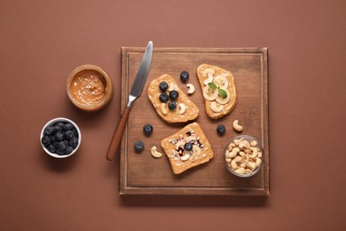 Photo of Toasts with tasty nut butter, blueberries, banana slices and cashews on brown table, flat lay