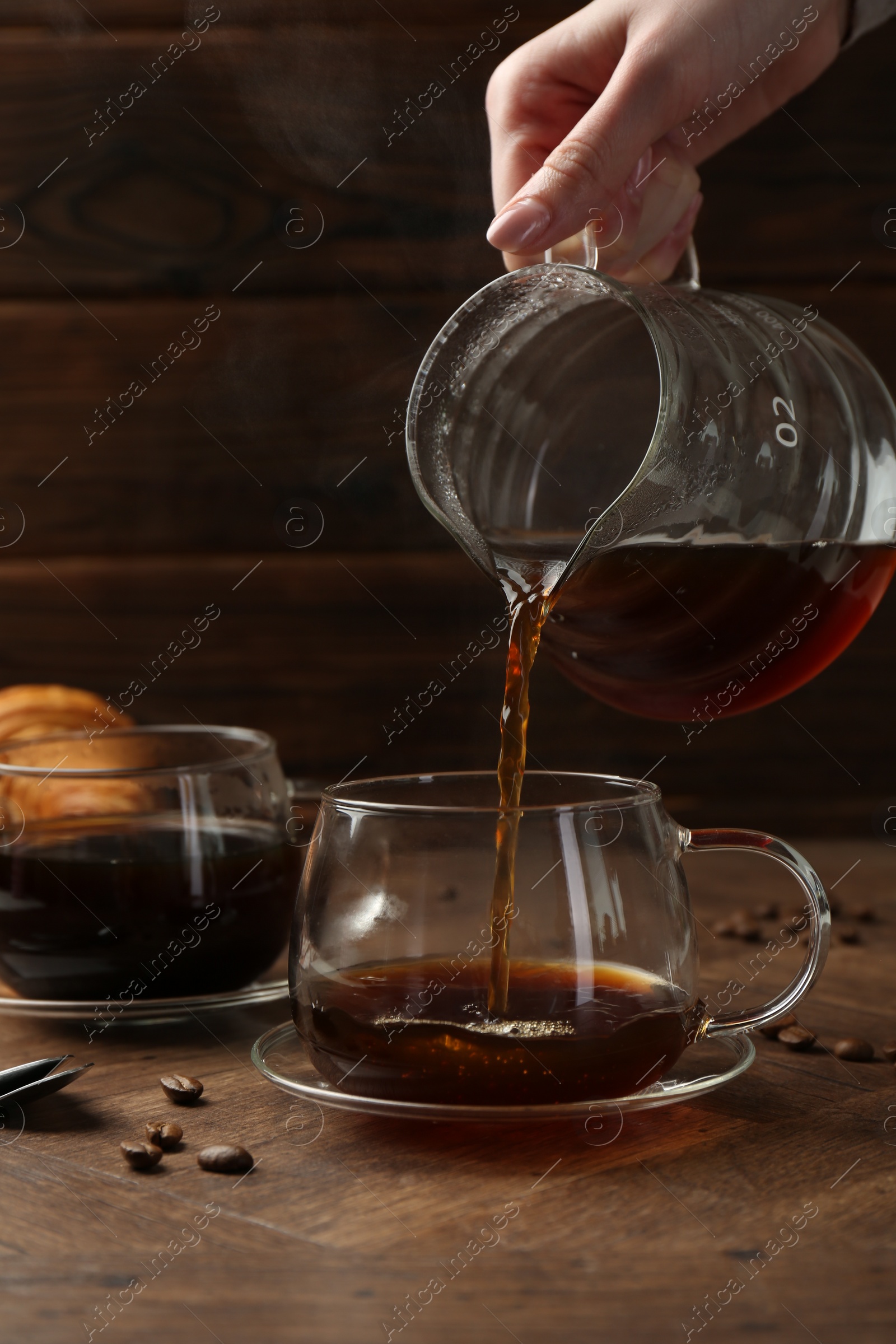 Photo of Woman pouring coffee into glass cup at wooden table, closeup