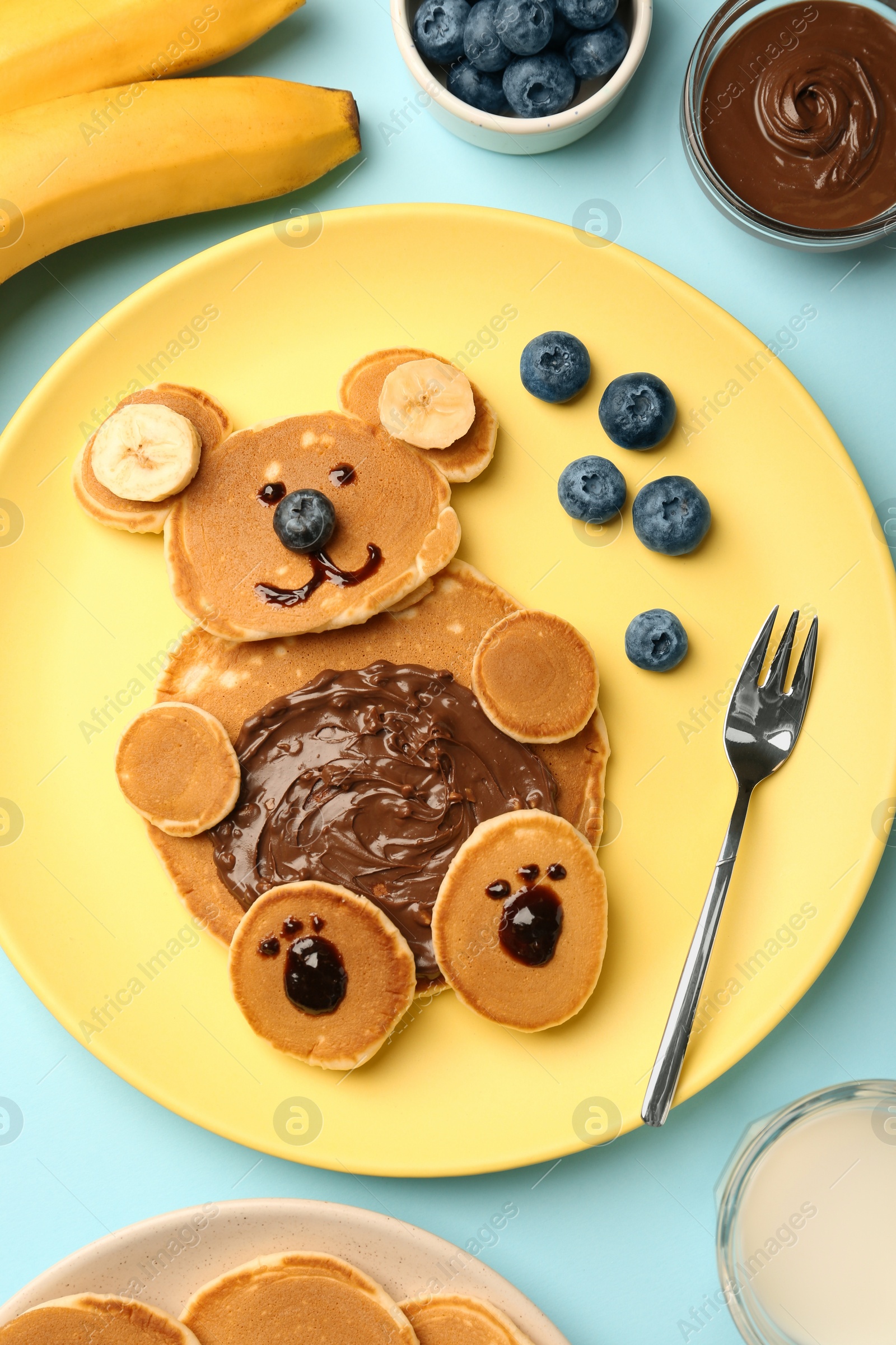 Photo of Creative serving for kids. Plate with cute bear made of pancakes, blueberries, bananas and chocolate paste on light blue table, flat lay