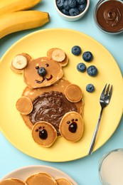 Creative serving for kids. Plate with cute bear made of pancakes, blueberries, bananas and chocolate paste on light blue table, flat lay