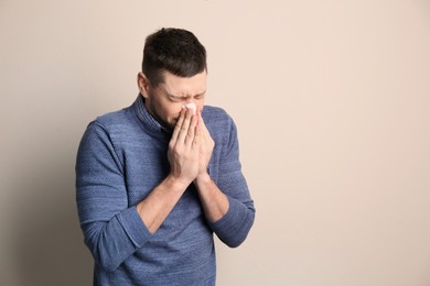 Man with tissue suffering from runny nose on beige background. Space for text