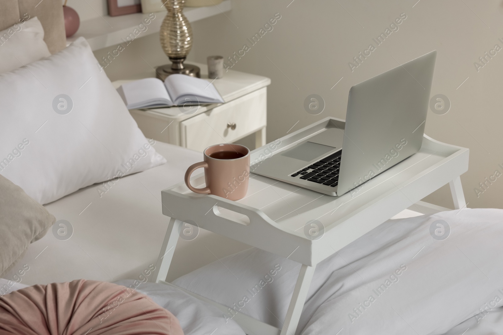 Photo of White tray table with laptop and cup of drink on bed indoors