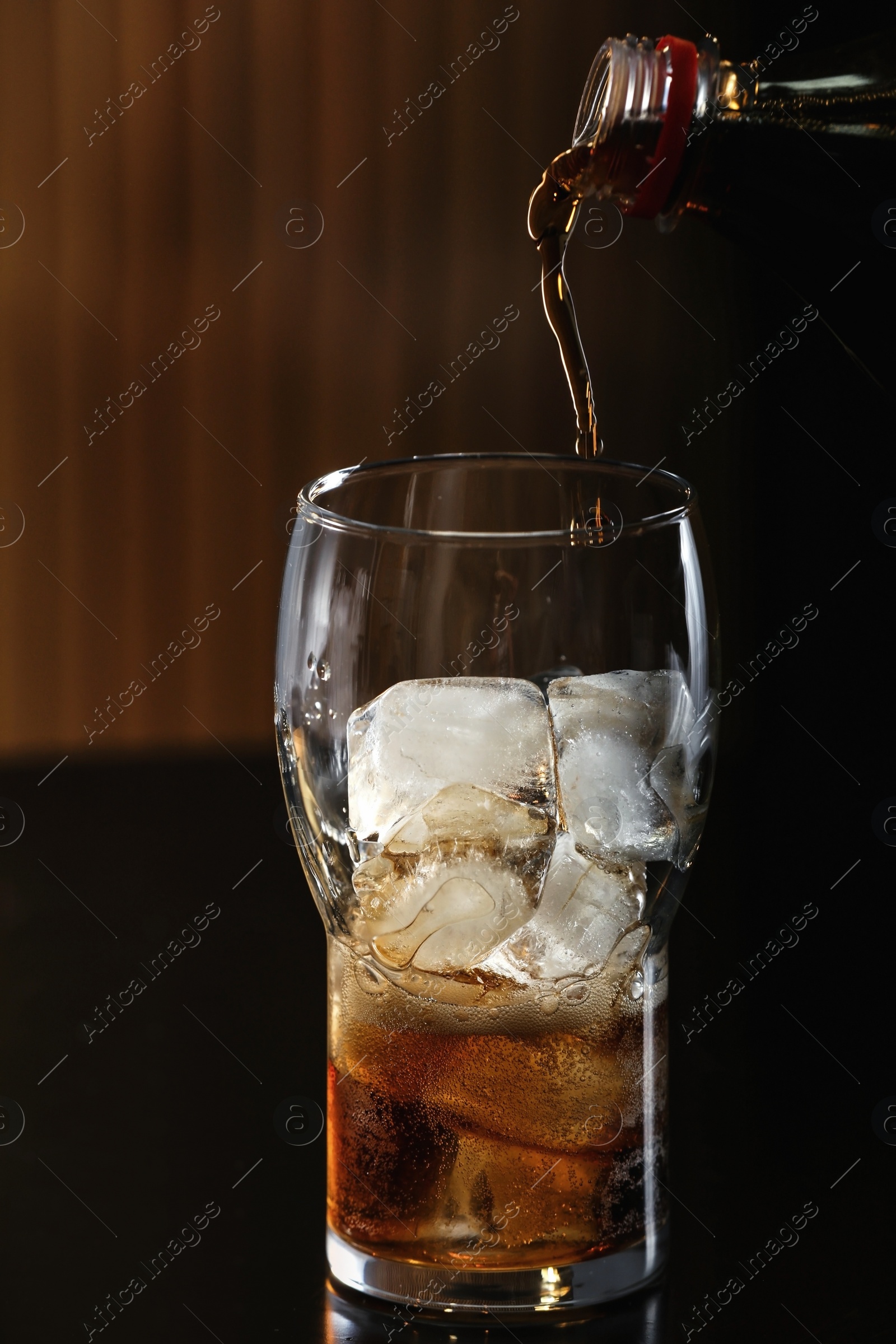 Photo of Pouring cola from bottle into glass with ice cubes on table against blurred background