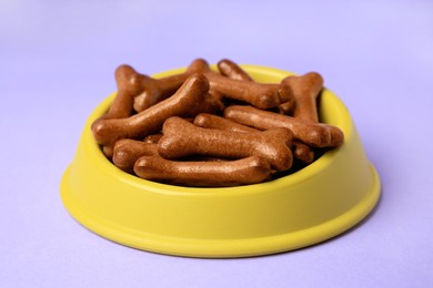 Photo of Yellow bowl with bone shaped dog cookies on purple background, closeup