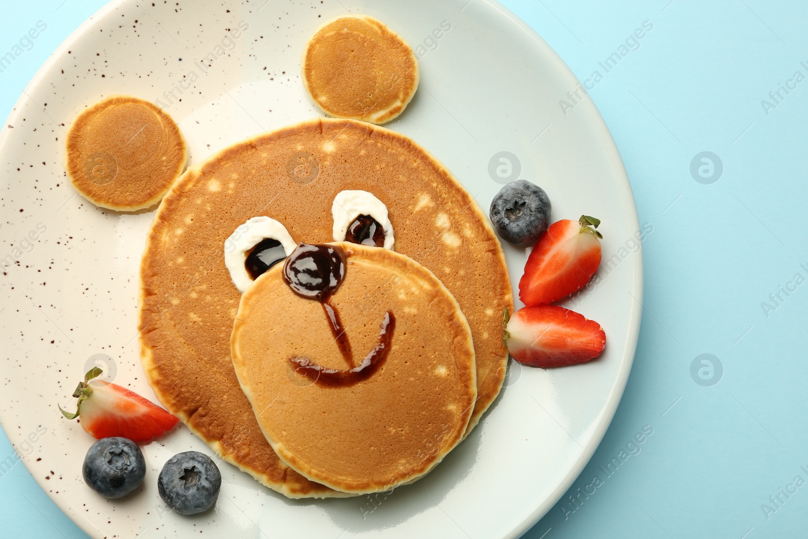 Photo of Creative serving for kids. Plate with cute bear made of pancakes and berries on light blue table, top view