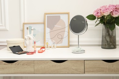 Photo of Mirror, cosmetic products, perfumes and vase with pink roses on white dressing table in makeup room