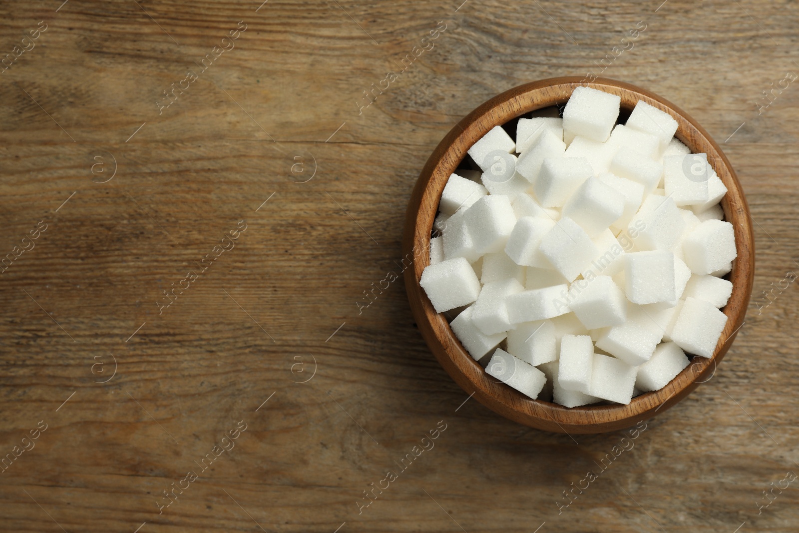 Photo of Bowl of white sugar cubes on wooden table, top view. Space for text