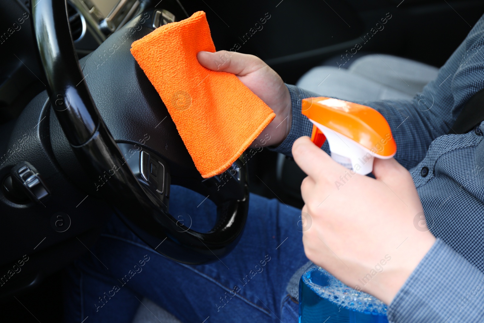 Photo of Man cleaning steering wheel with rag and spray bottle in car, closeup