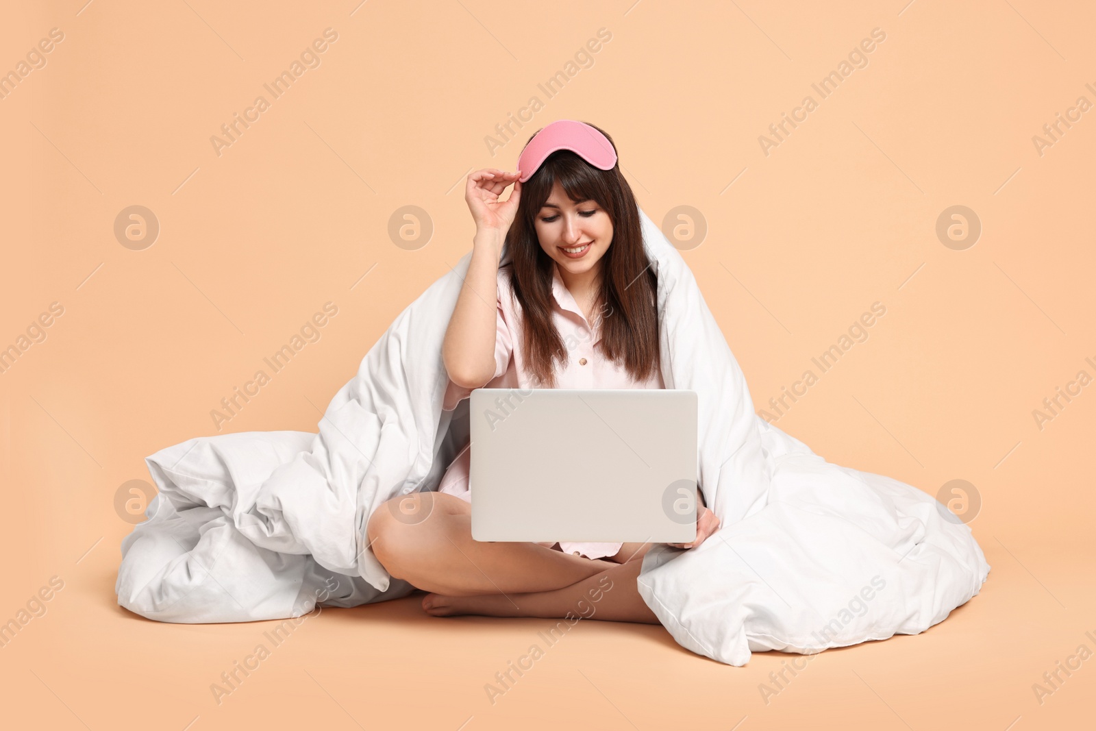 Photo of Happy woman with pyjama and blanket holding laptop on beige background