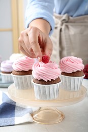 Woman decorating delicious cupcakes with fresh raspberries at table indoors, closeup