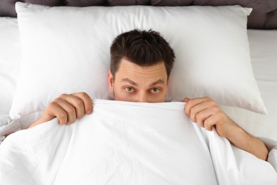 Handsome man covering his face with blanket while lying on pillow, top view. Bedtime