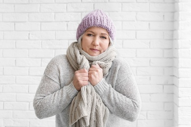 Mature woman in warm clothes suffering from cold on brick background