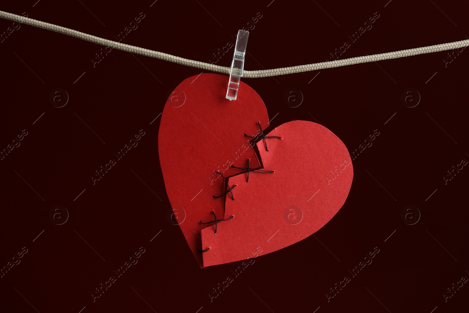 Photo of Broken heart. Torn red paper heart sewed with thread on rope against burgundy background