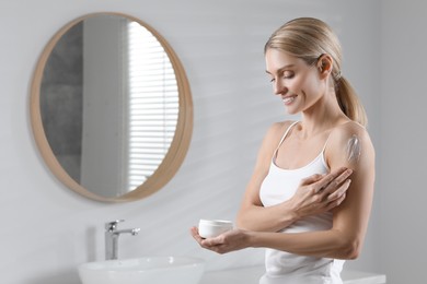 Photo of Happy woman applying body cream onto arm in bathroom, space for text