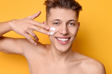 Photo of Handsome man applying moisturizing cream onto his face on orange background