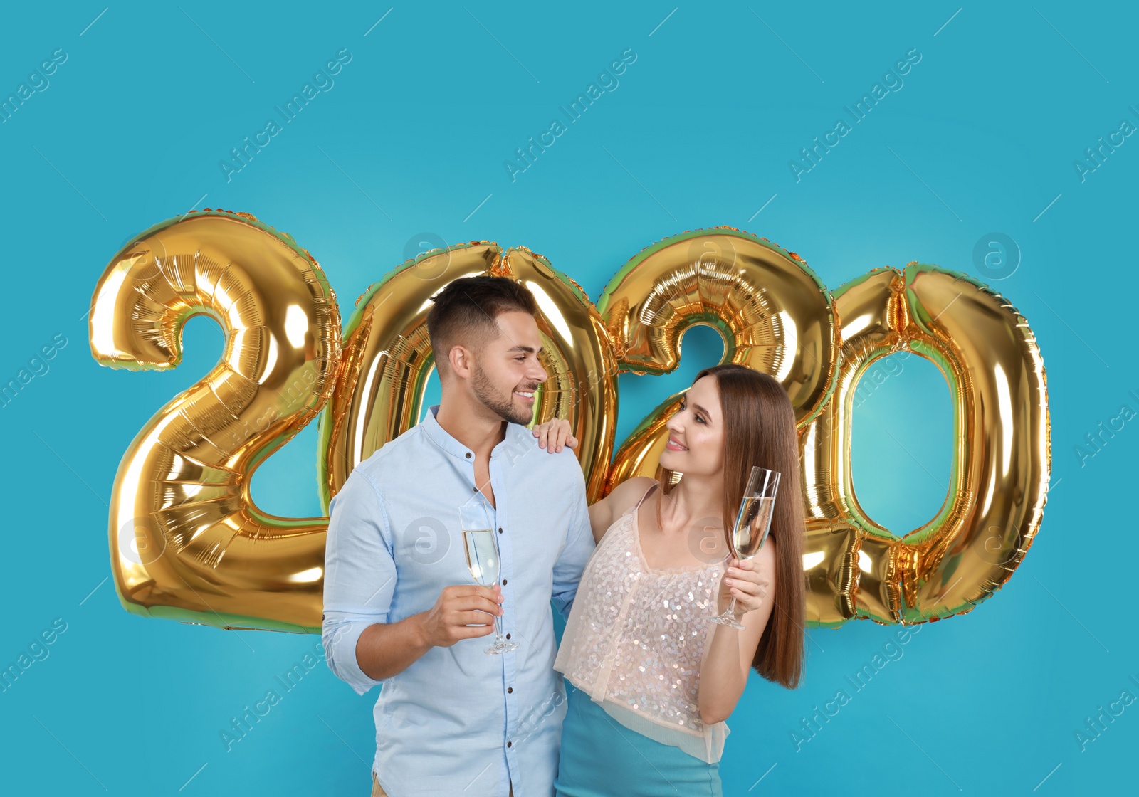 Photo of Happy young couple with glasses of champagne near golden 2020 balloons on turquoise background. New Year celebration