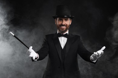 Photo of Happy magician holding wand in smoke on black background