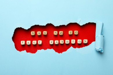 Photo of Phrase SAY NO TO FAST FASHION made of wooden cubes on red background, view through hole in light blue paper, flat lay