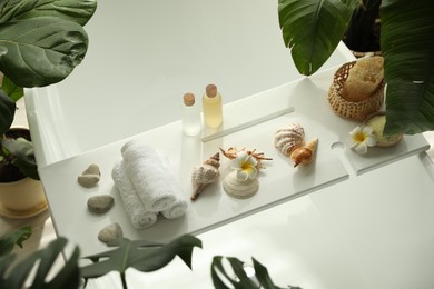 Bath tray with spa products, towels and shells on tub in bathroom, above view