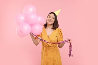 Happy young woman in party hat with balloons on pink background