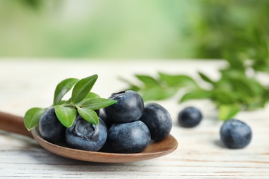 Wooden spoon of fresh blueberries with leaves on white table against blurred green background, space for text